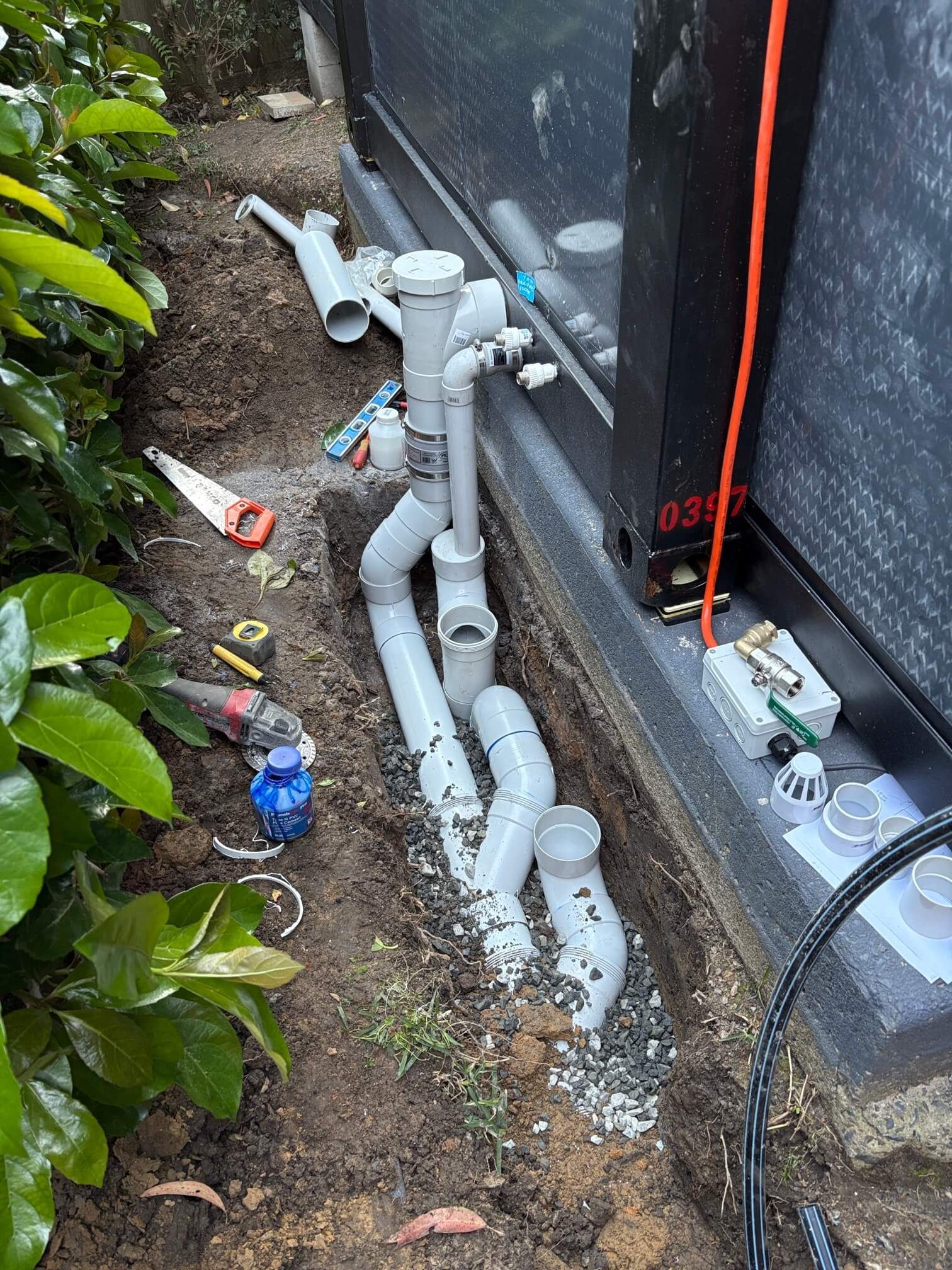 Gray PVC plumbing pipes installed in a narrow trench next to a dark building wall, surrounded by soil and tools.