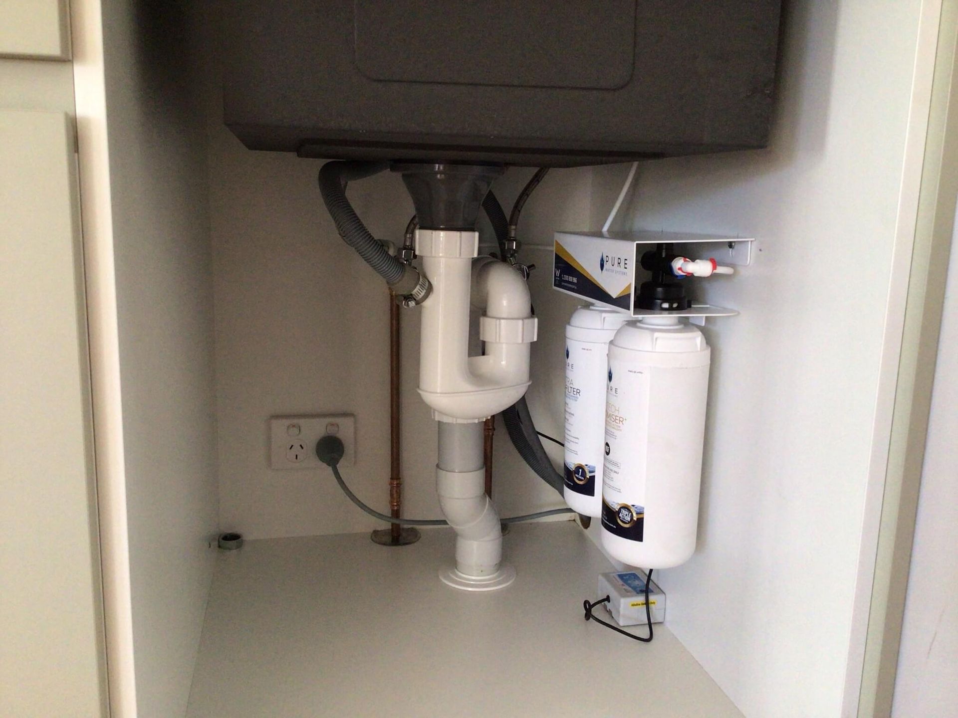 Under-sink area showing white water filtration system units mounted on a side wall next to white PVC plumbing pipes.