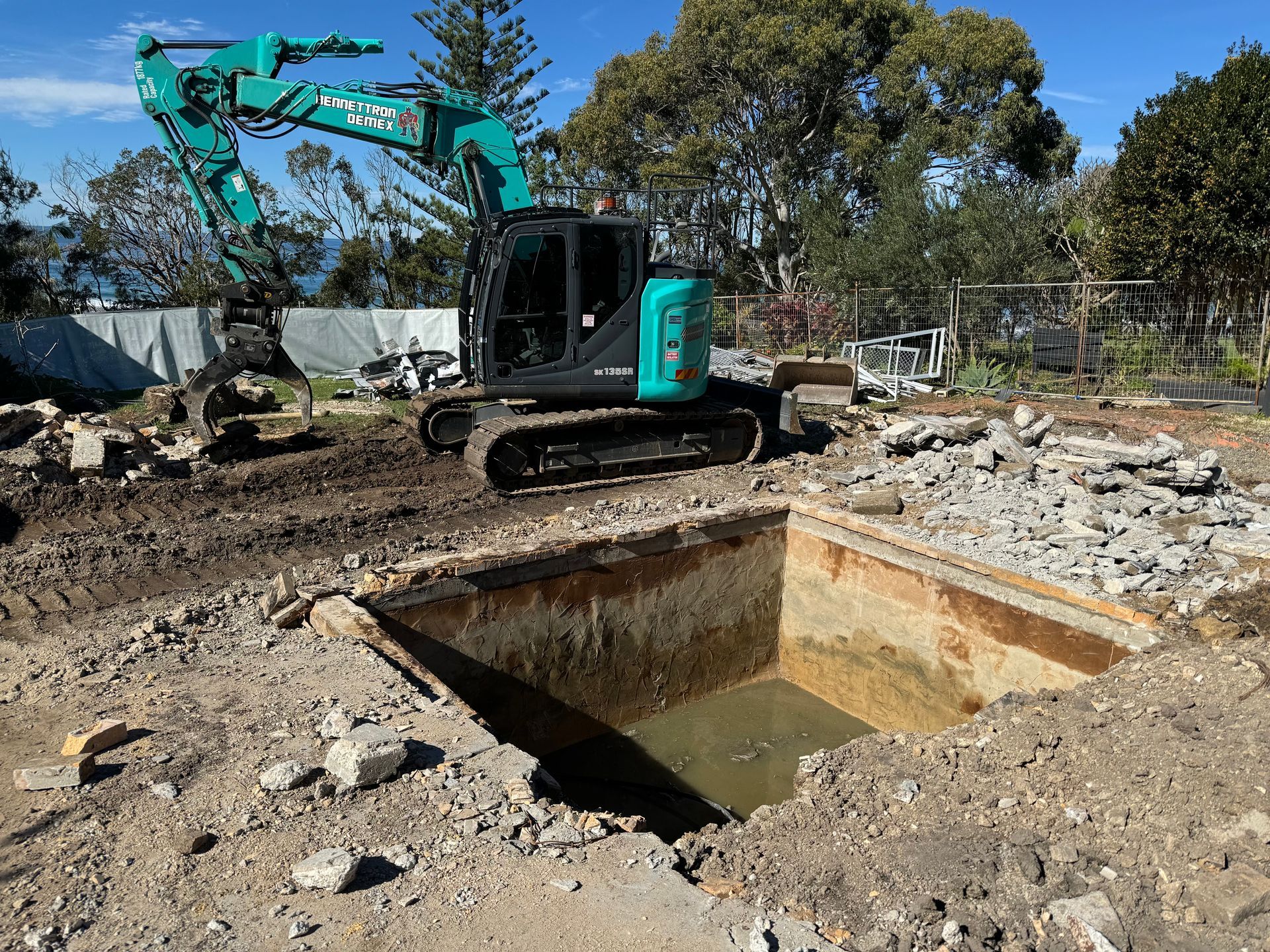 A Large Square hole has been dug in dirt lot with blue digger parked beside it — Bennettron Demex in Corindi Beach, NSW