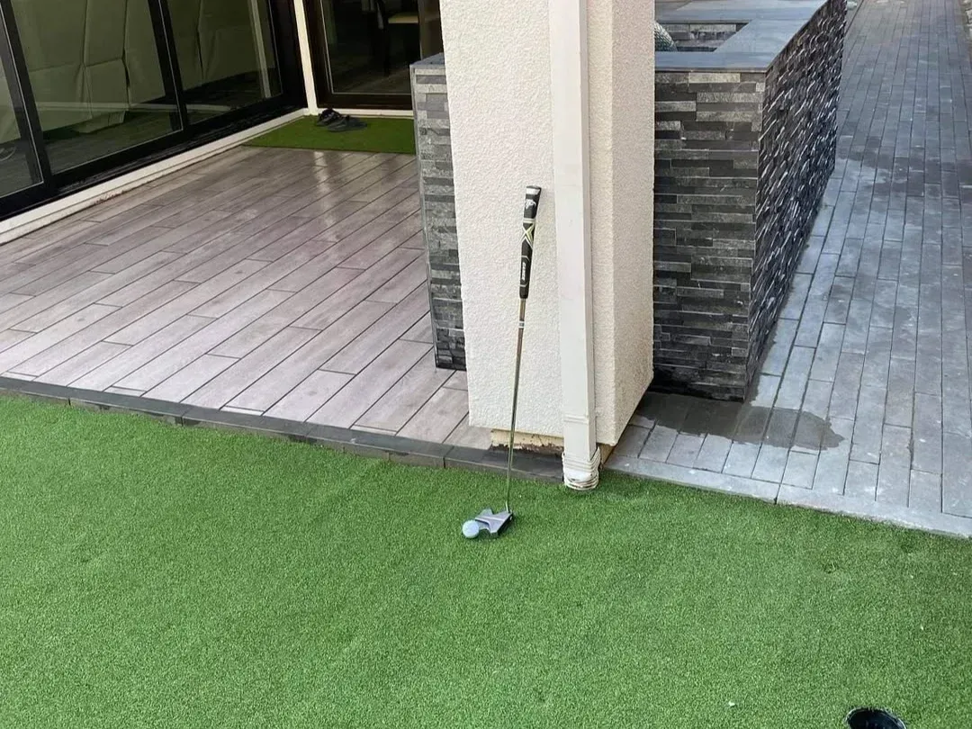 Patio corner with artificial grass, wooden deck, tiled walkway, and a broom leaning against a wall