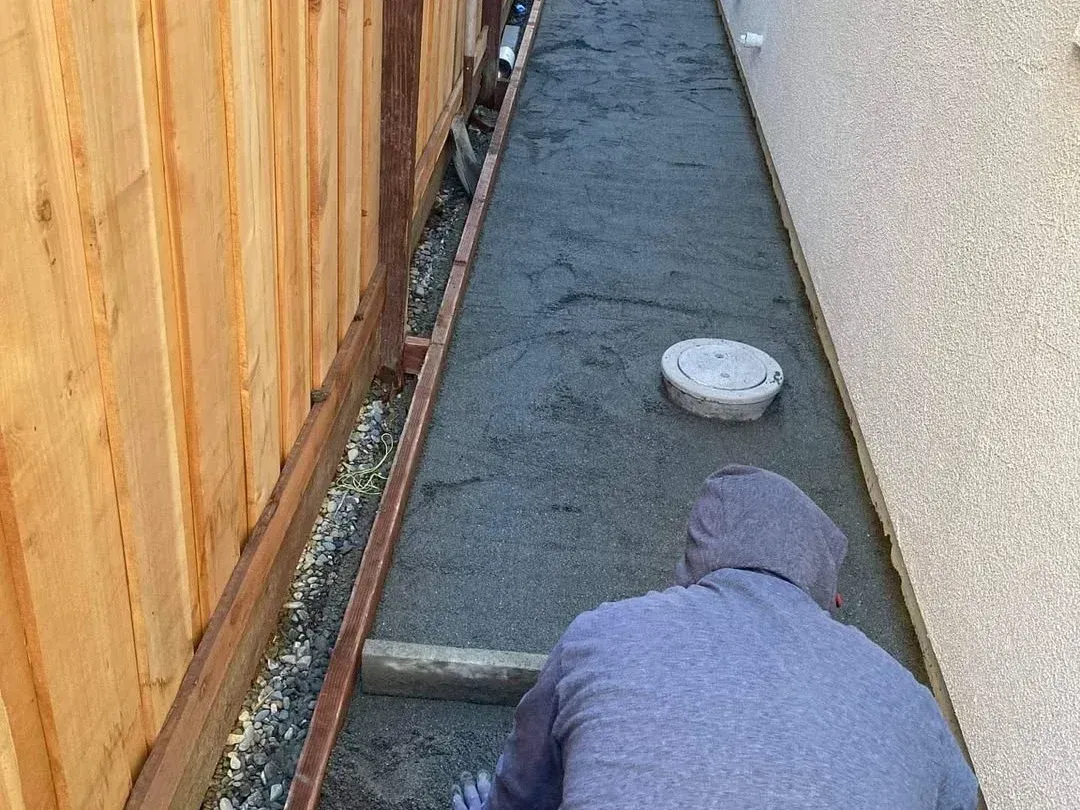 Worker in a narrow trench applying concrete beside a wooden fence and house wall.