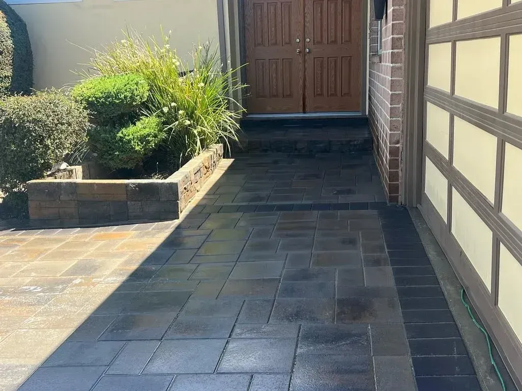 Paved walkway leading to a wooden front door beside a house and garden bed