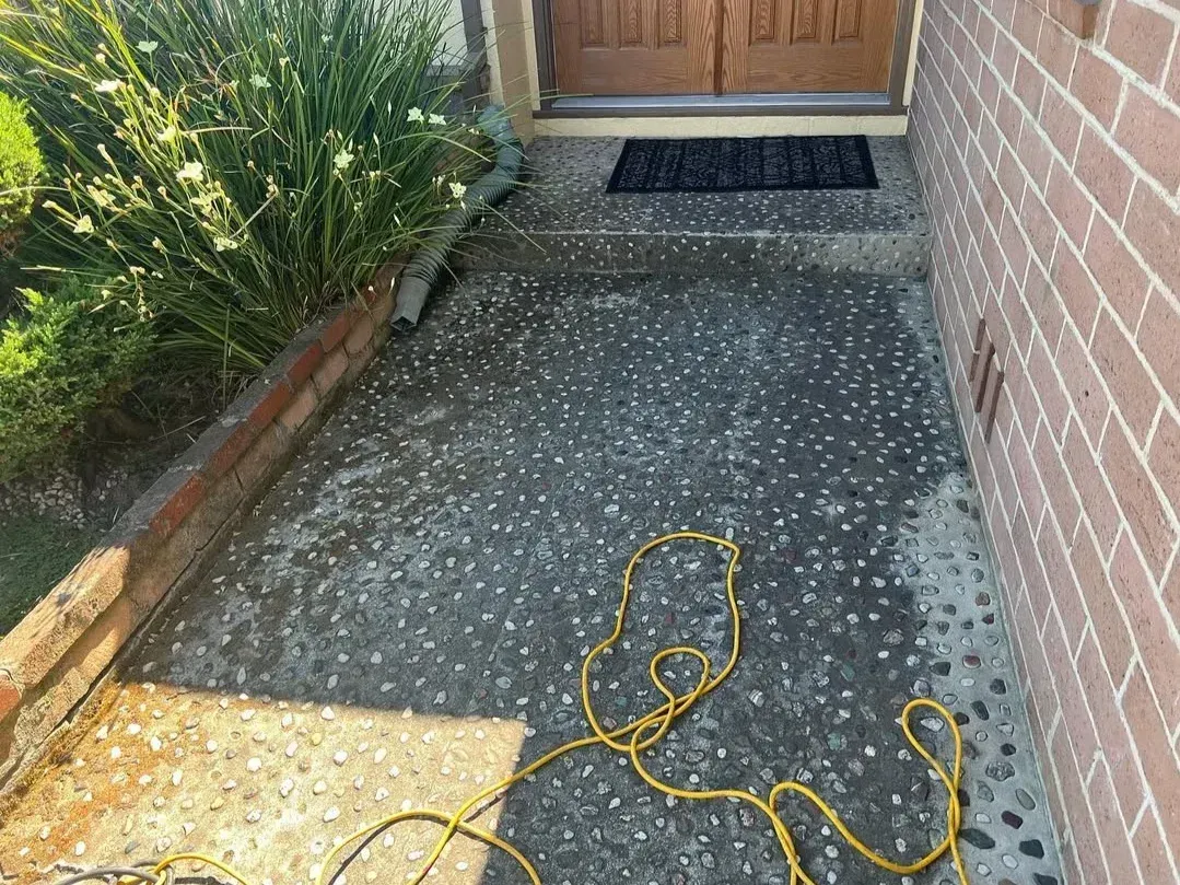 Narrow concrete walkway by brick wall and garden, with a coiled yellow cord on the ground near a wooden door