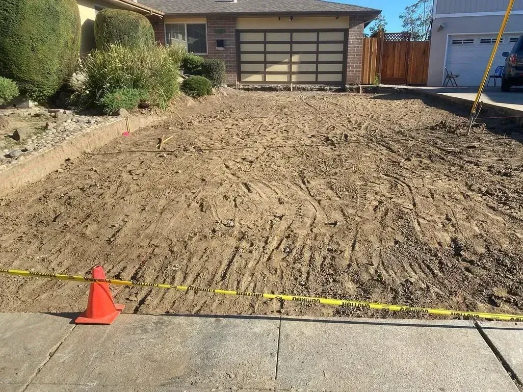 Front yard cleared to bare dirt beside a garage, with orange cones and caution tape at the curb.