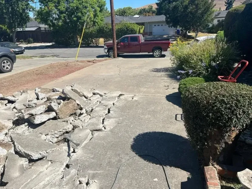 Damaged concrete walkway with broken rocks beside a suburban driveway and parked red pickup truck.