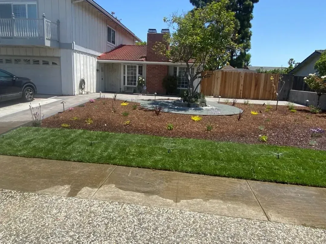 Suburban front yard with a driveway, garage, lawn, and mulched garden beds in bright sunlight