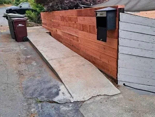 Concrete sidewalk beside a red wooden fence with a black mailbox and red trash bin on a driveway.