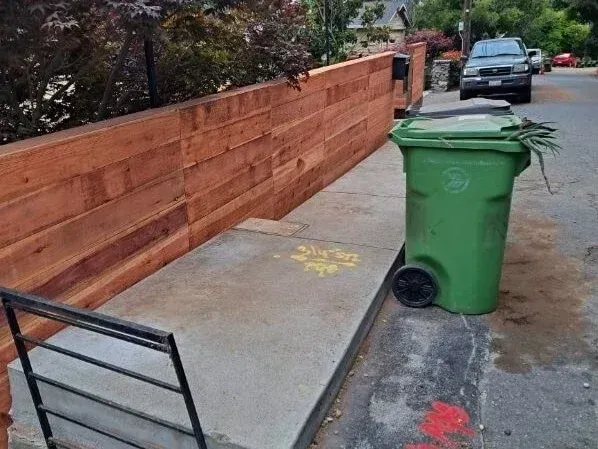 Sidewalk with a green trash bin blocking the curb lane beside a brick wall.