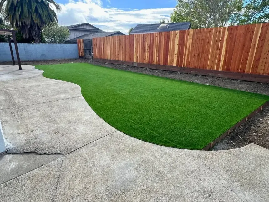 Backyard with bright green lawn, concrete patio, and tall wooden fence under a blue sky