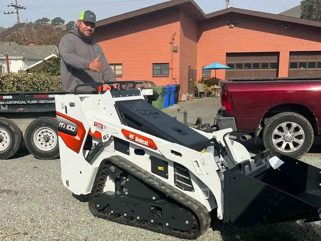 Man standing on a white Bobcat skid steer beside a red pickup in a gravel lot