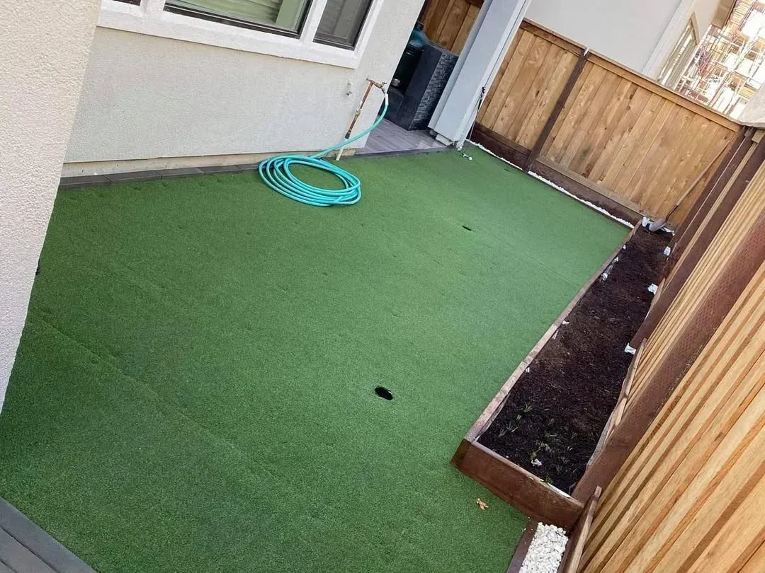 Small backyard with artificial grass, wooden fence, raised planter, and a coiled blue hose near the house.