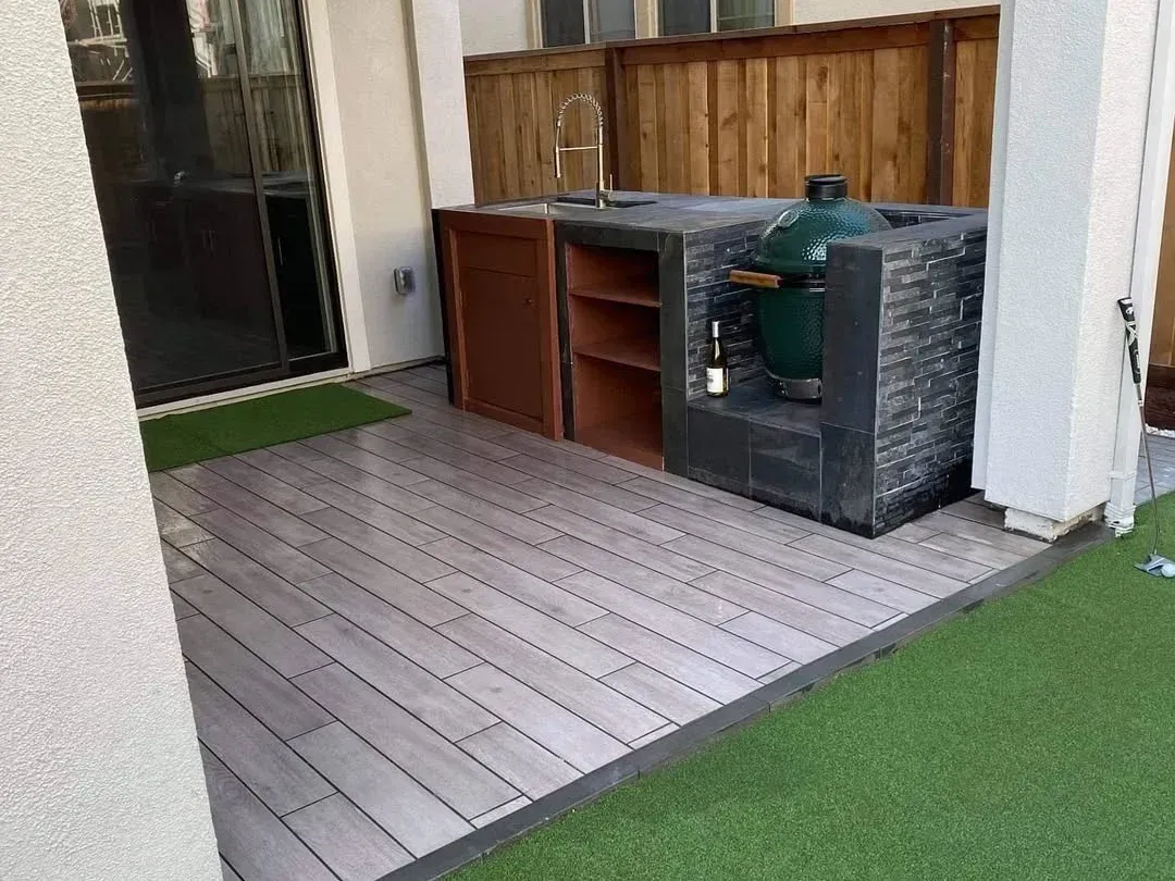 Outdoor patio with gray deck tiles, artificial grass, wooden cabinets, and a grill against the wall