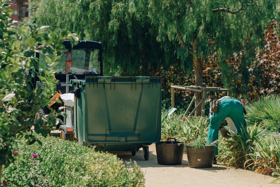 Person in green working beside a large green dumpster in a lush garden area.
