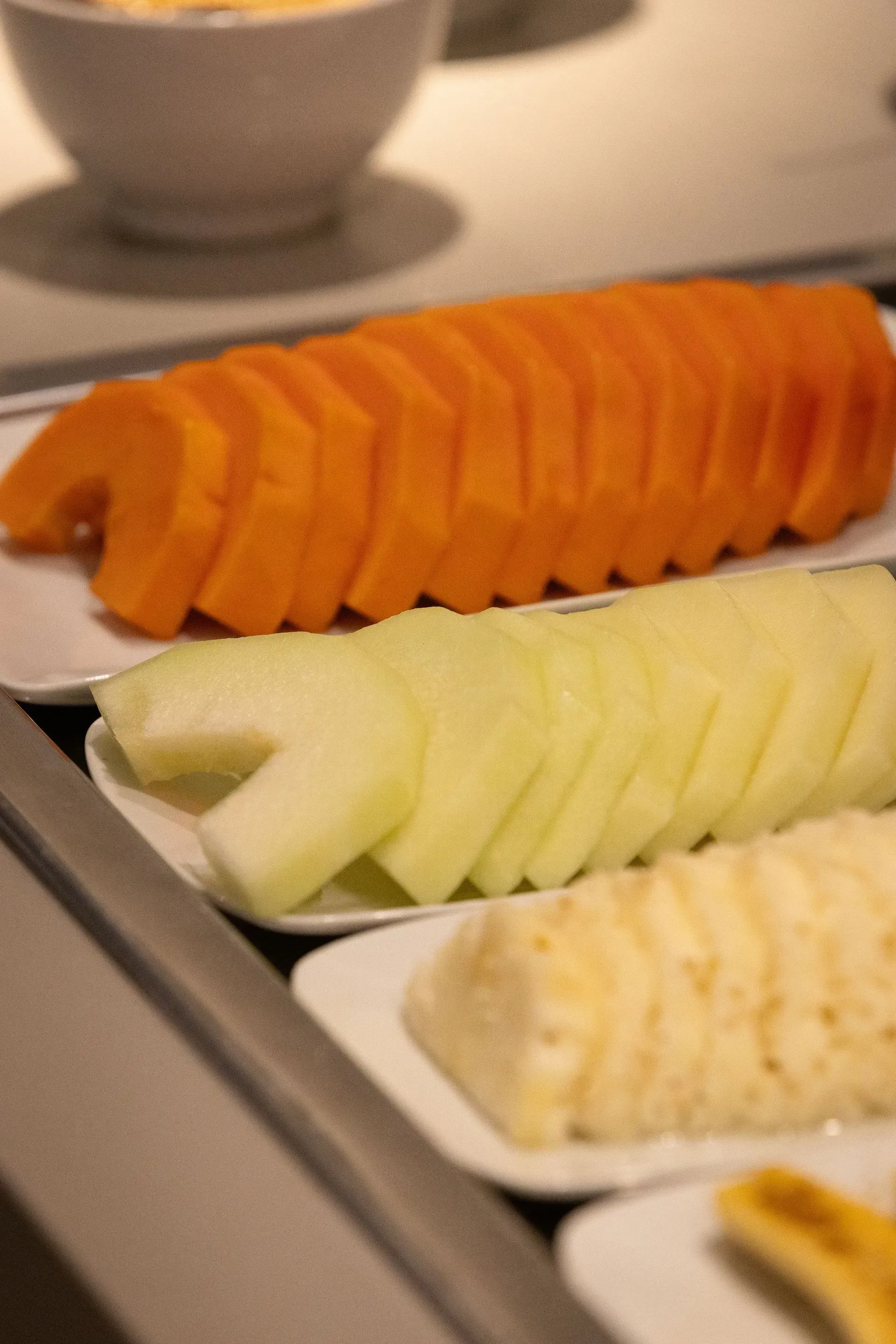 Fatias de mamão laranja, melão verde e frutas amarelas em pratos brancos, dispostas em um balcão.