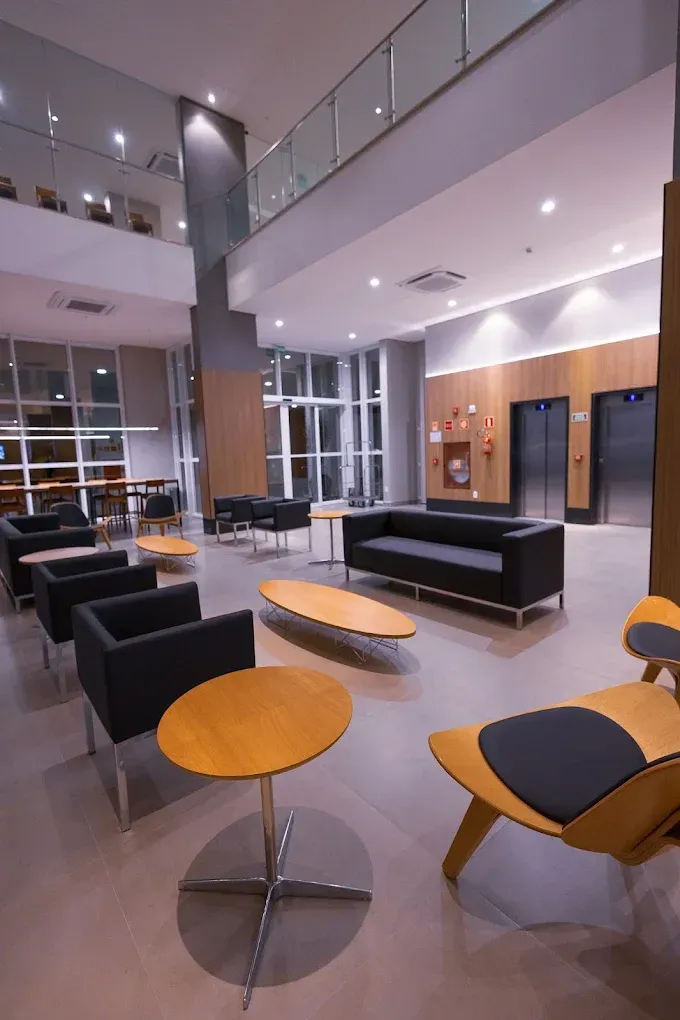 Hall de entrada com áreas de estar: sofás, poltronas e mesas redondas. Elevador e grandes janelas ao fundo.