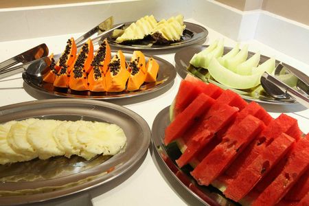 Um bufê de frutas com travessas de prata contendo fatias de melancia, abacaxi, mamão e melão em uma bancada branca.