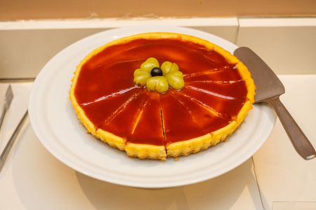 Uma tarte fatiada com um glacê vermelho brilhante, coberta com uvas verdes, servida num prato branco com uma espátula.