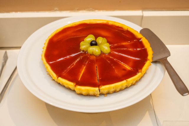 Uma tarte fatiada com um glacê vermelho brilhante, coberta com uvas verdes, servida num prato branco com uma espátula.