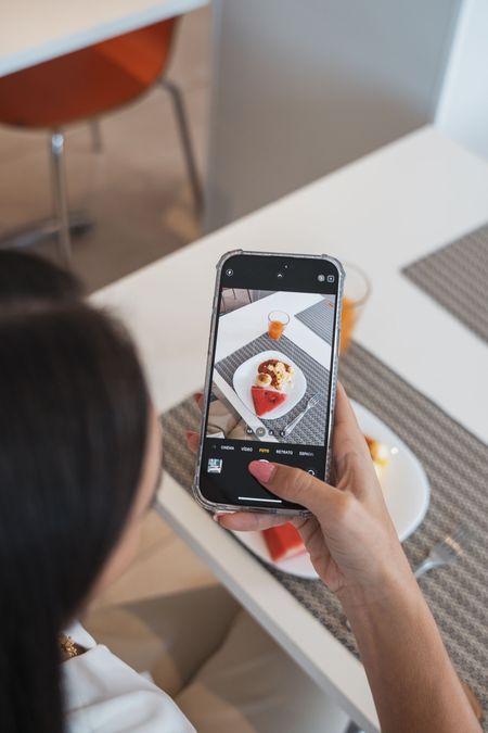 Uma pessoa segura um celular para tirar uma foto de um pequeno doce em formato de coração sobre um jogo americano estampado.