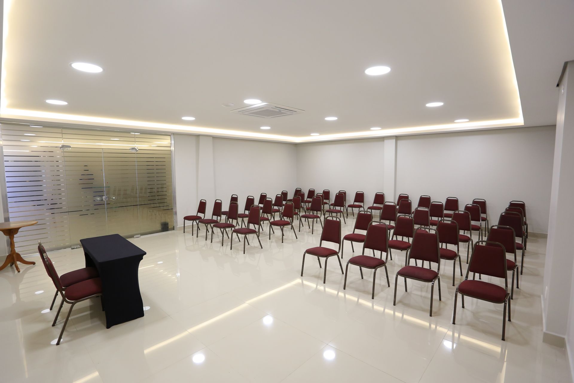 Sala de eventos com fileiras de mesas cobertas com toalhas, cadeiras azuis, paredes em tons neutros e iluminação no teto.