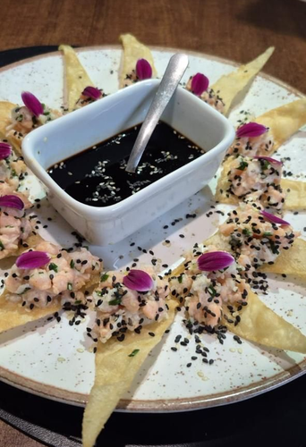 Prato de wonton fritos em formato triangular, cobertos com  salmão, sementes de gergelim e flores roxas, com molho de soja.