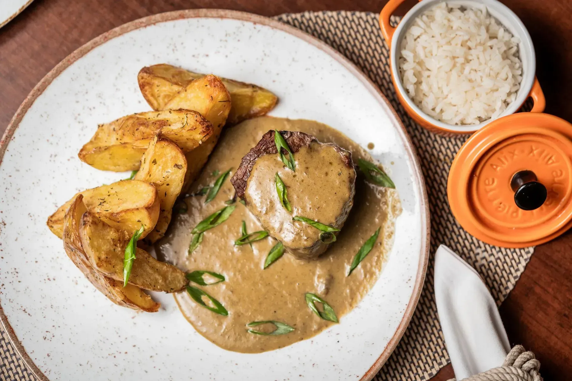 Bife com molho e batatas rústicas em um prato, arroz em uma tigela, tampa de laranja.