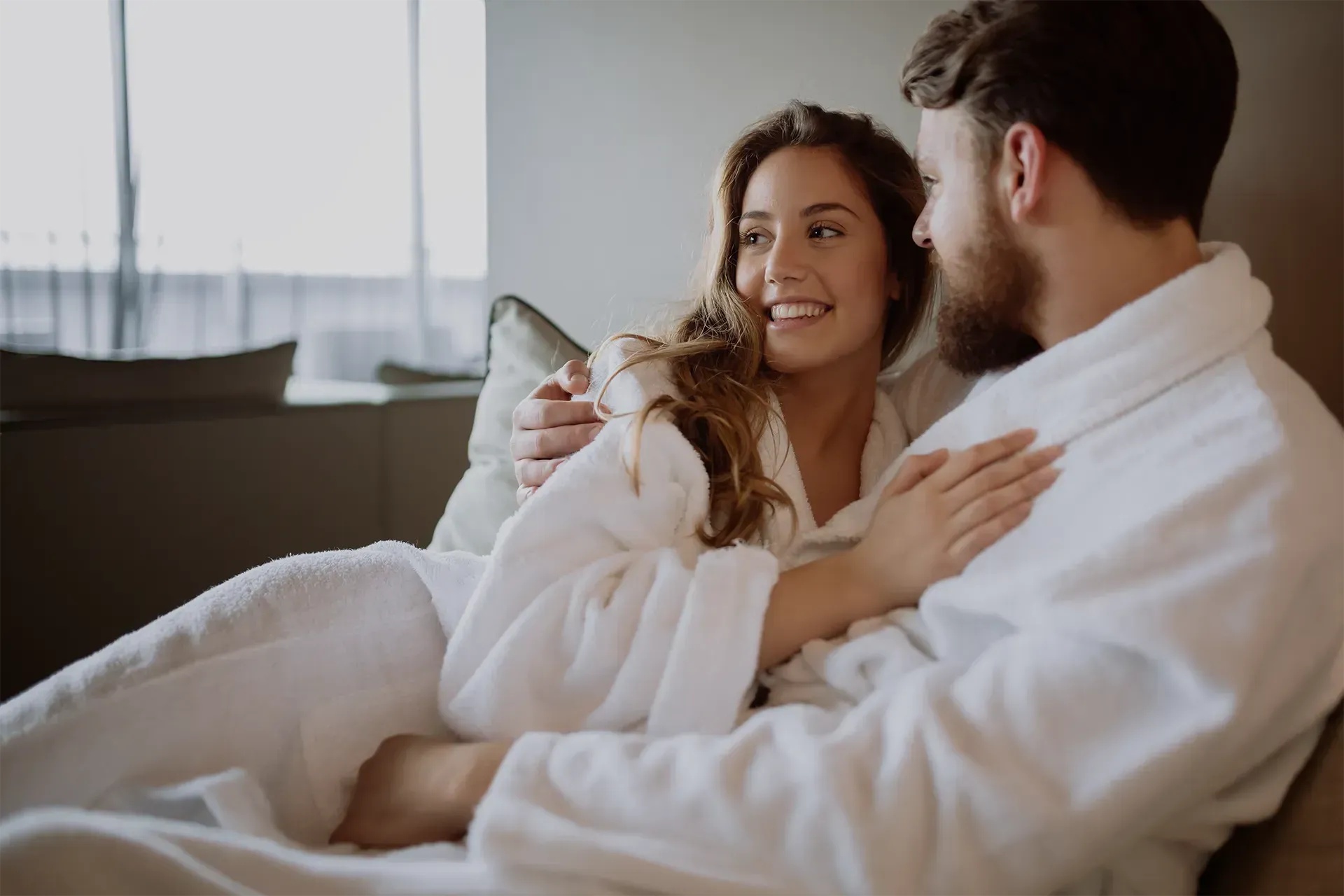 Um casal de roupões brancos, sorrindo e se abraçando em uma cama de quarto de hotel.