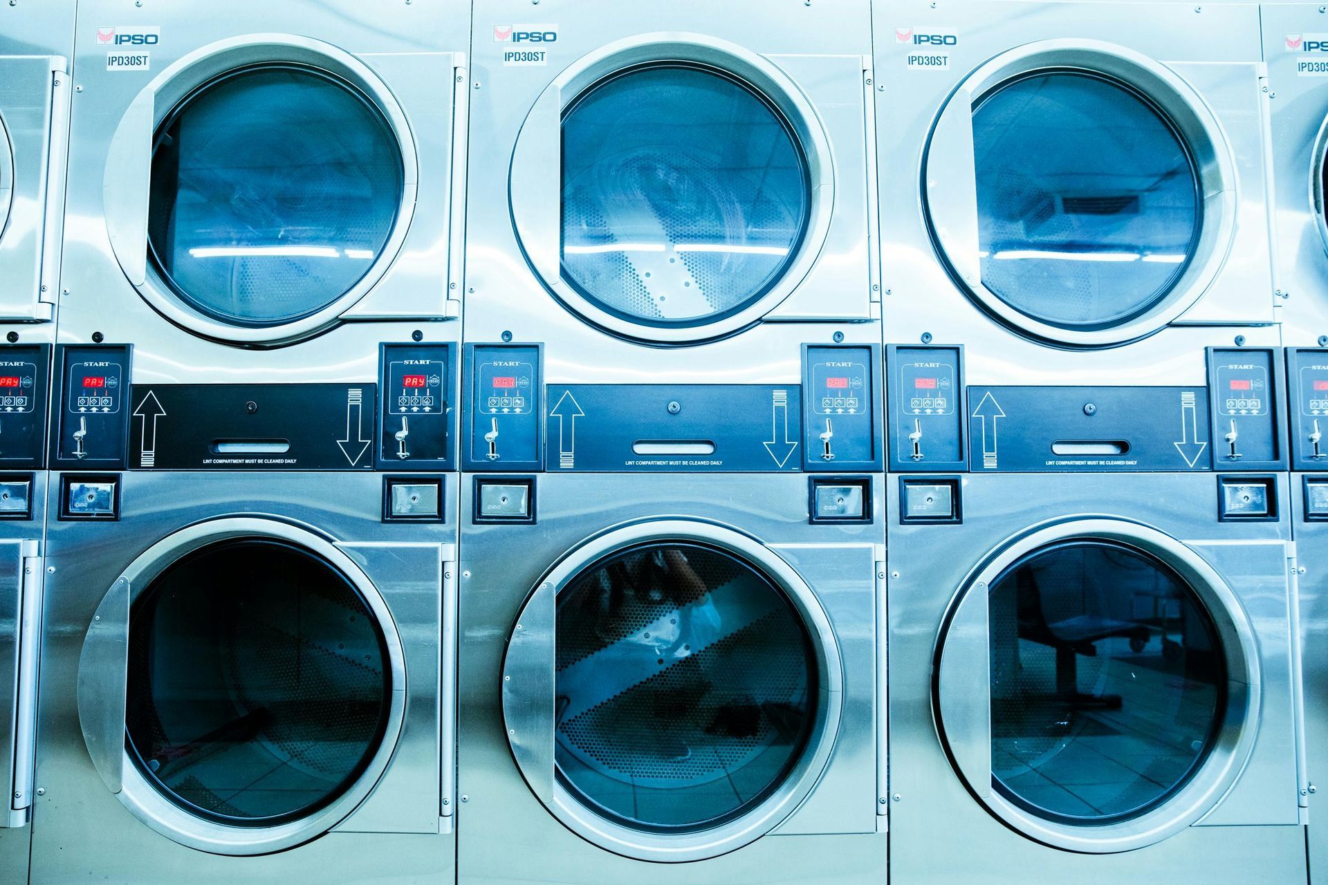 Row of stainless steel commercial clothes dryers in a laundromat, with glass doors and control panels.