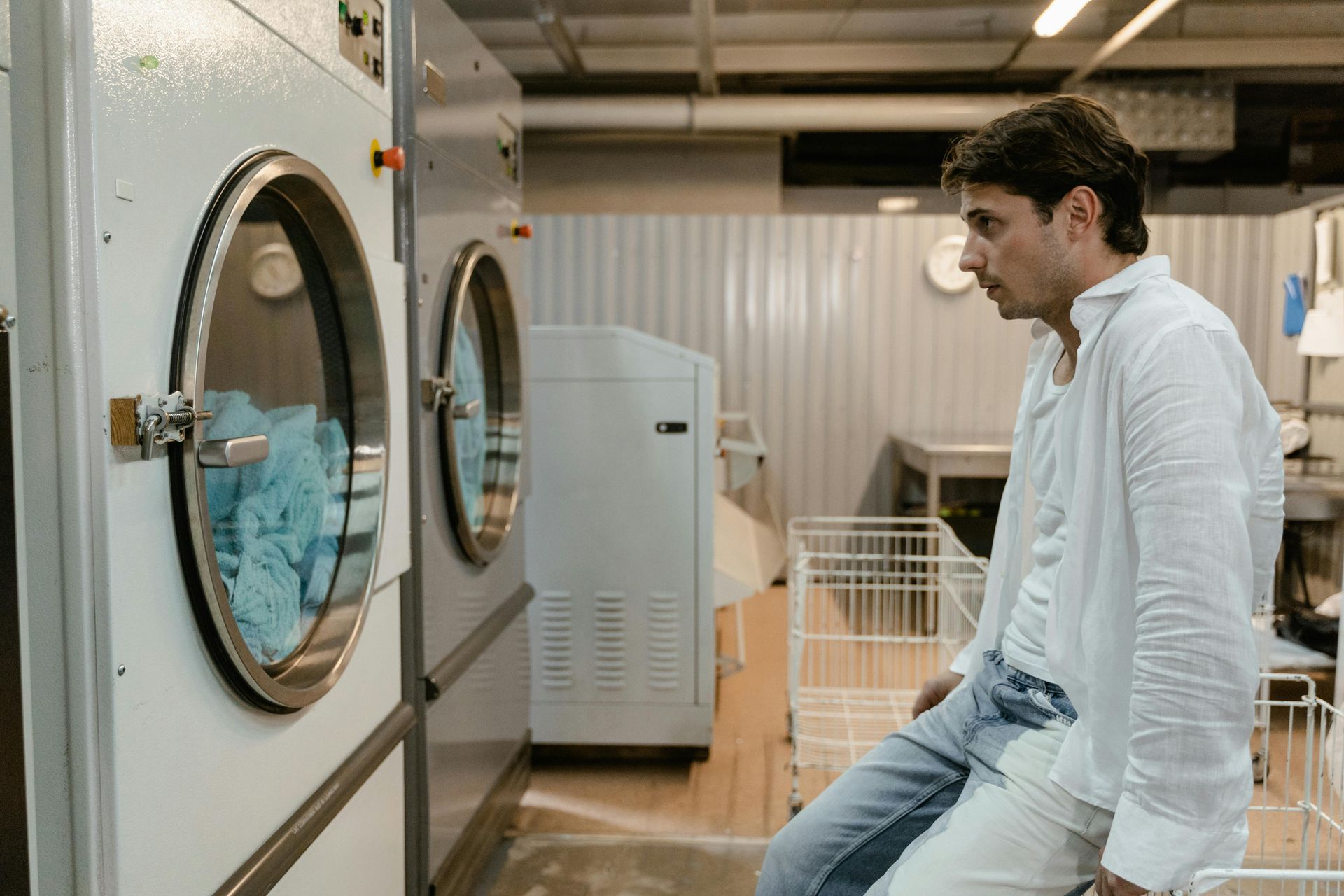 Man looking into a dryer at a laundromat. He’s wearing a white shirt and leaning on the machine.