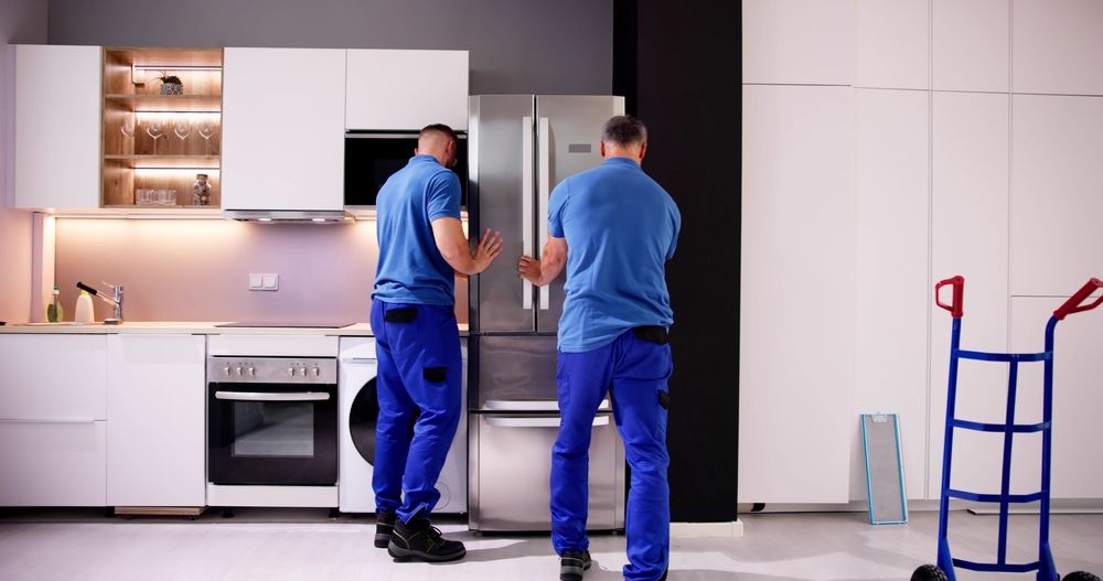 Two movers in blue uniforms pushing a stainless steel refrigerator in a modern kitchen.