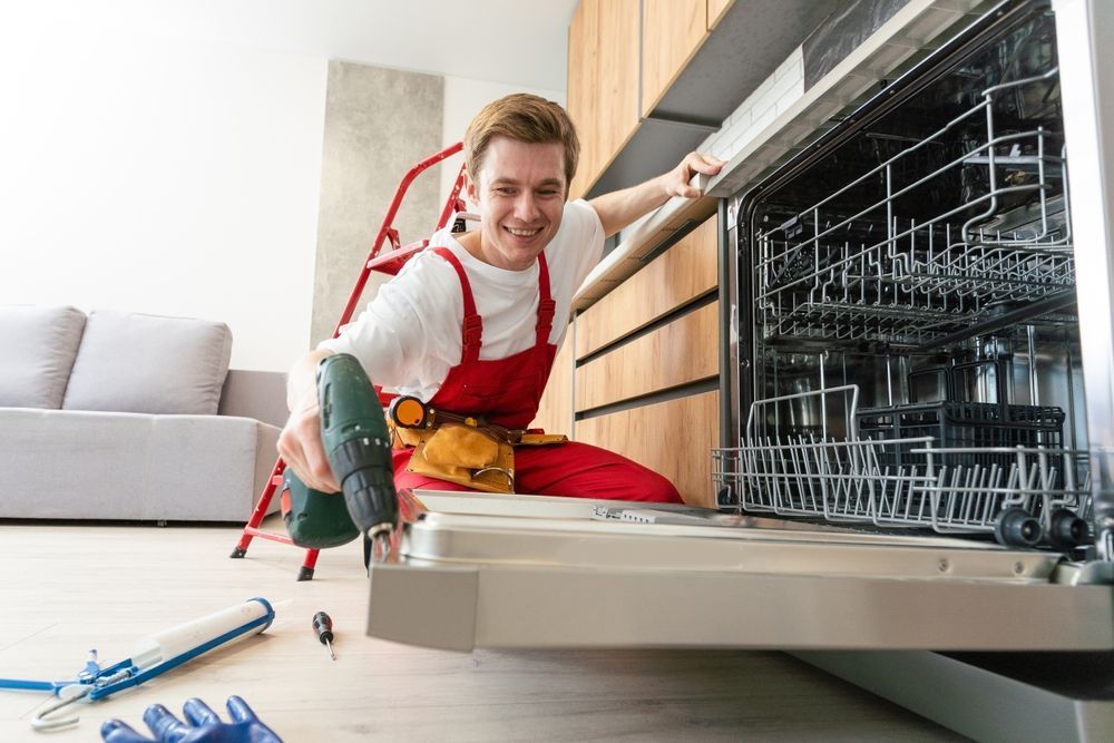 Man in red overalls repairing dishwasher with a drill.