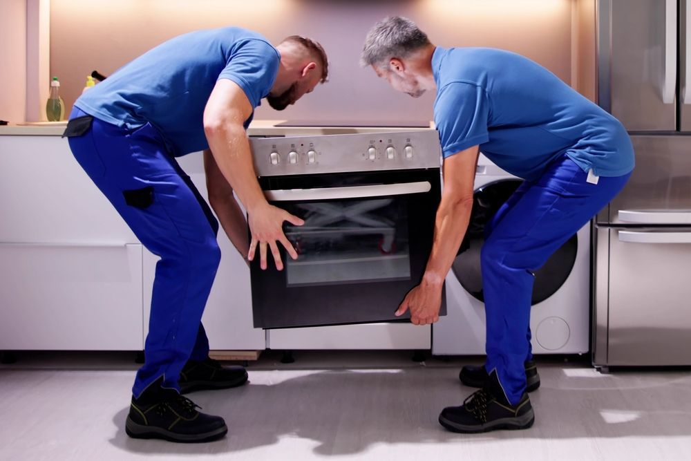 Two workers in blue uniforms lift an oven in a kitchen.
