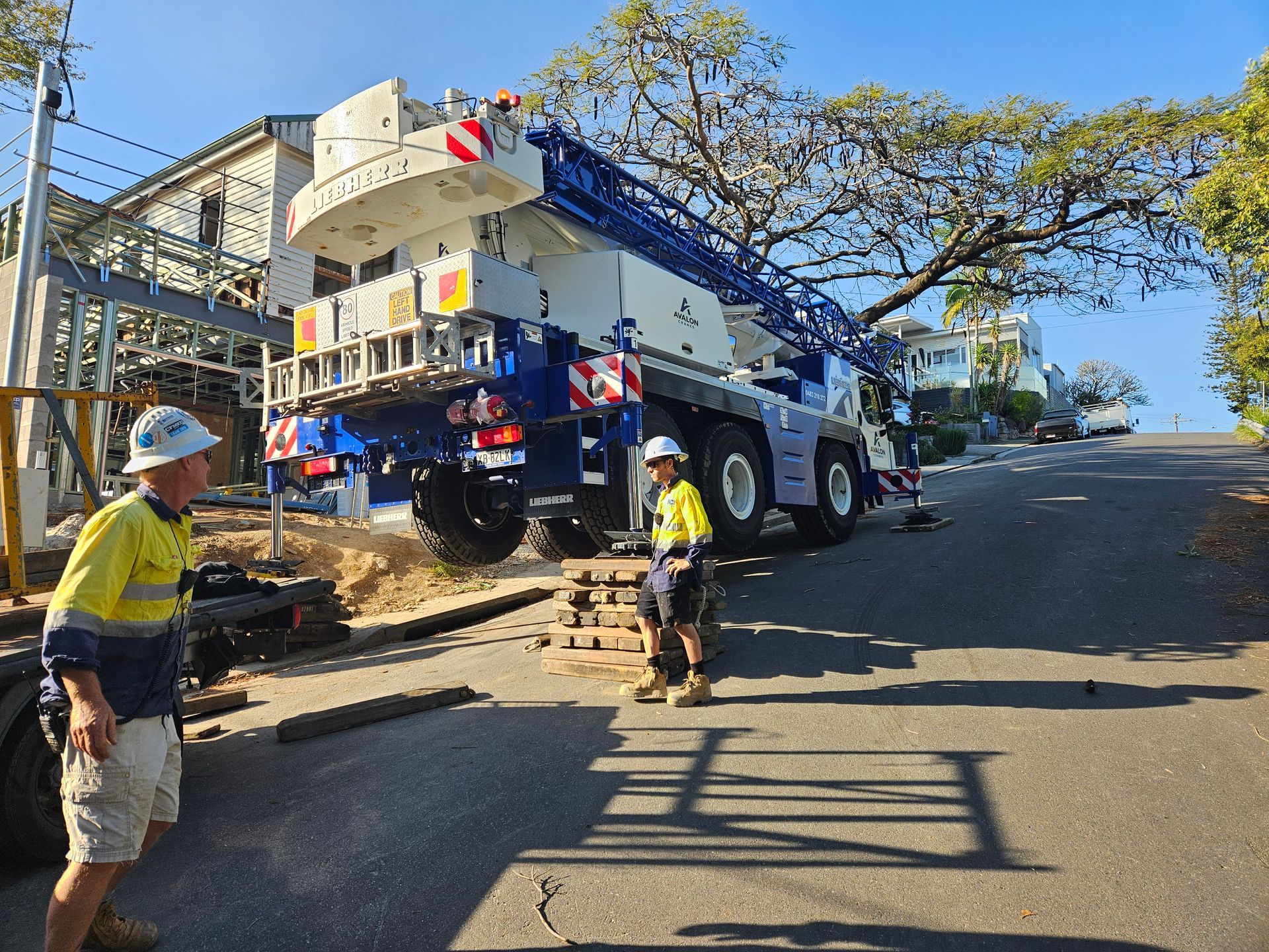 Workers guide a large crane on a steep street, near a building, and under a tree.