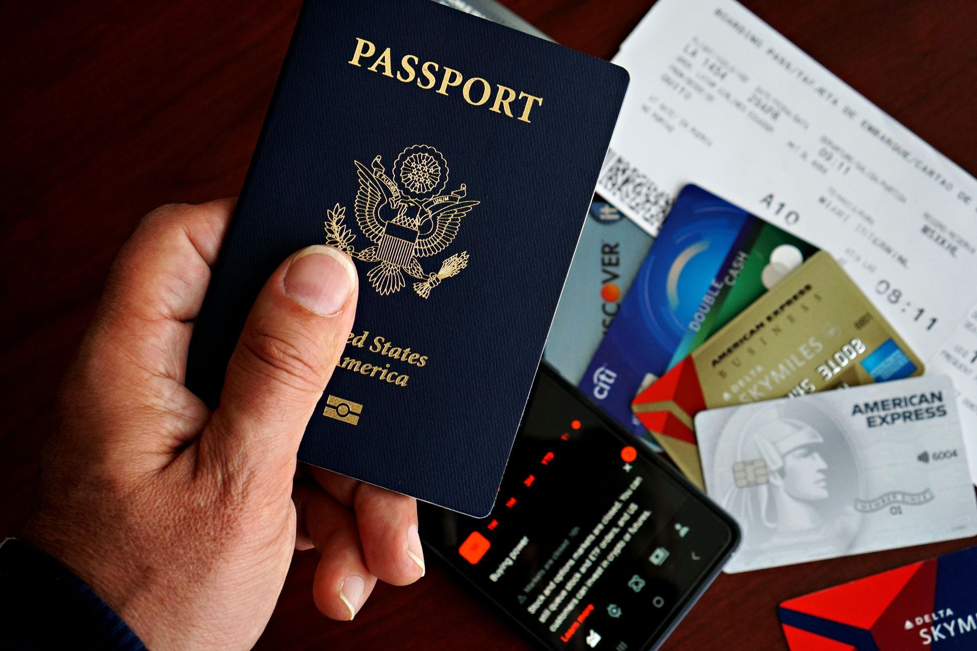 Hand holding a United States passport next to credit cards, a phone, and airline tickets.