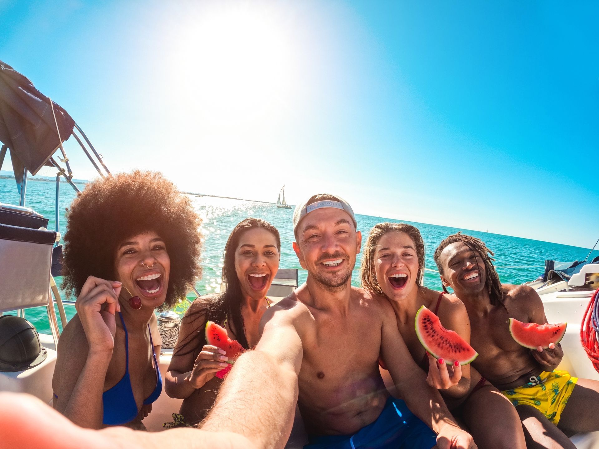 Group of friends on a boat selfie, holding watermelon, smiling, blue sky and ocean background.