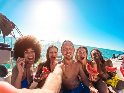 Group of friends on a boat, taking a selfie and eating watermelon under a bright blue sky and sun.