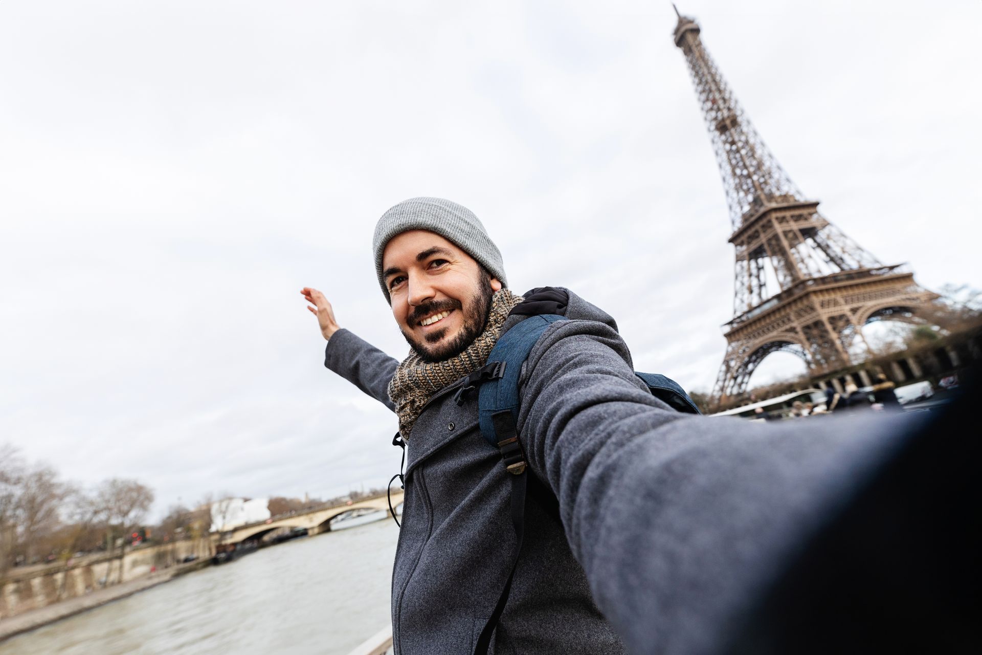 Man takes a selfie in Paris with the Eiffel Tower in the background, arms outstretched. Cloudy day.
