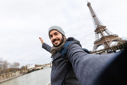 Man with a beard takes a selfie in front of the Eiffel Tower in Paris, France. He is smiling, wearing a hat, coat, and backpack.
