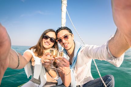 A couple on a sailboat taking a selfie, toasting with champagne. The setting is sunny with blue sky and water.