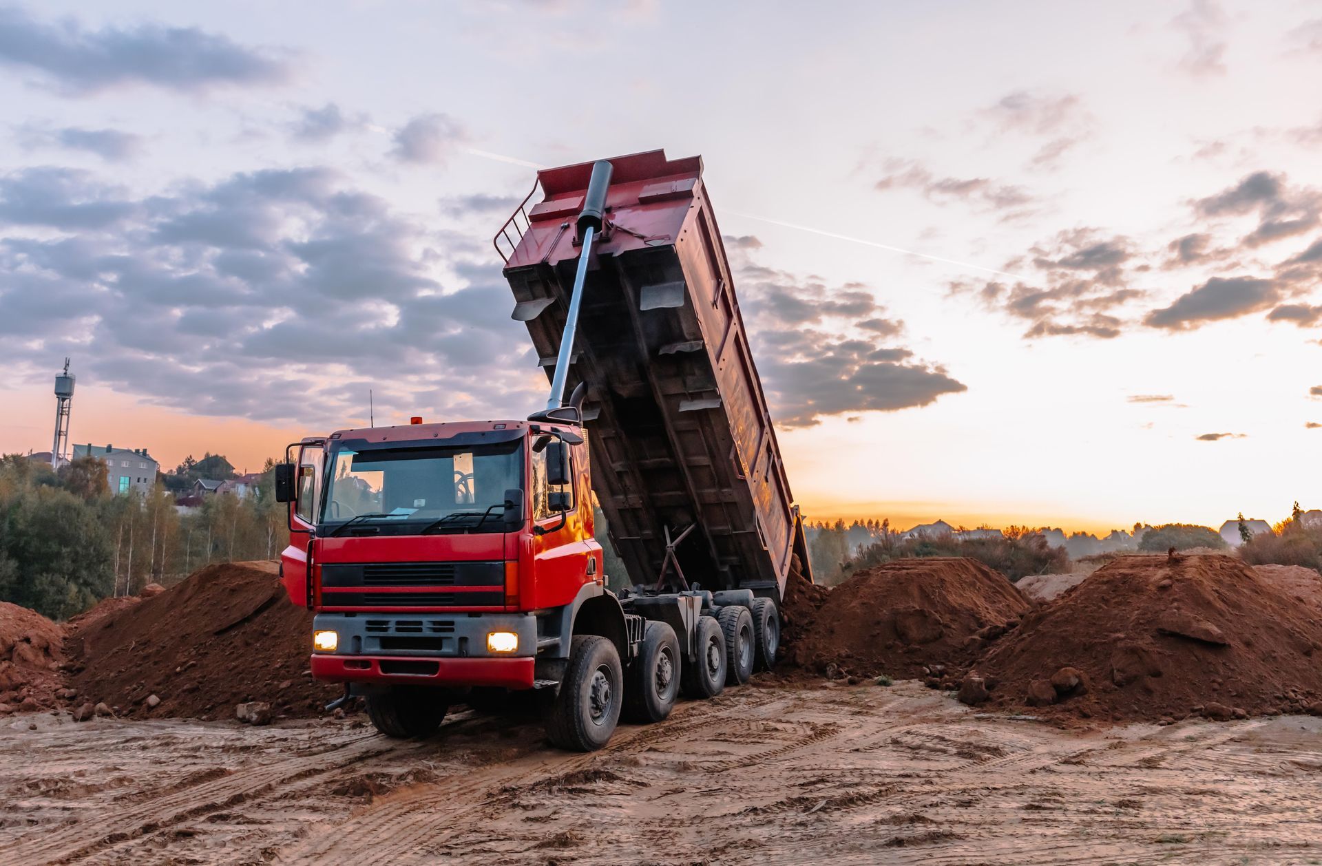 Red dump truck dumping soil onto a pile in a construction site at dusk.