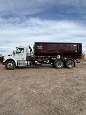 Photograph of a large white and maroon dump truck parked on a dirt surface under a partly cloudy sky.