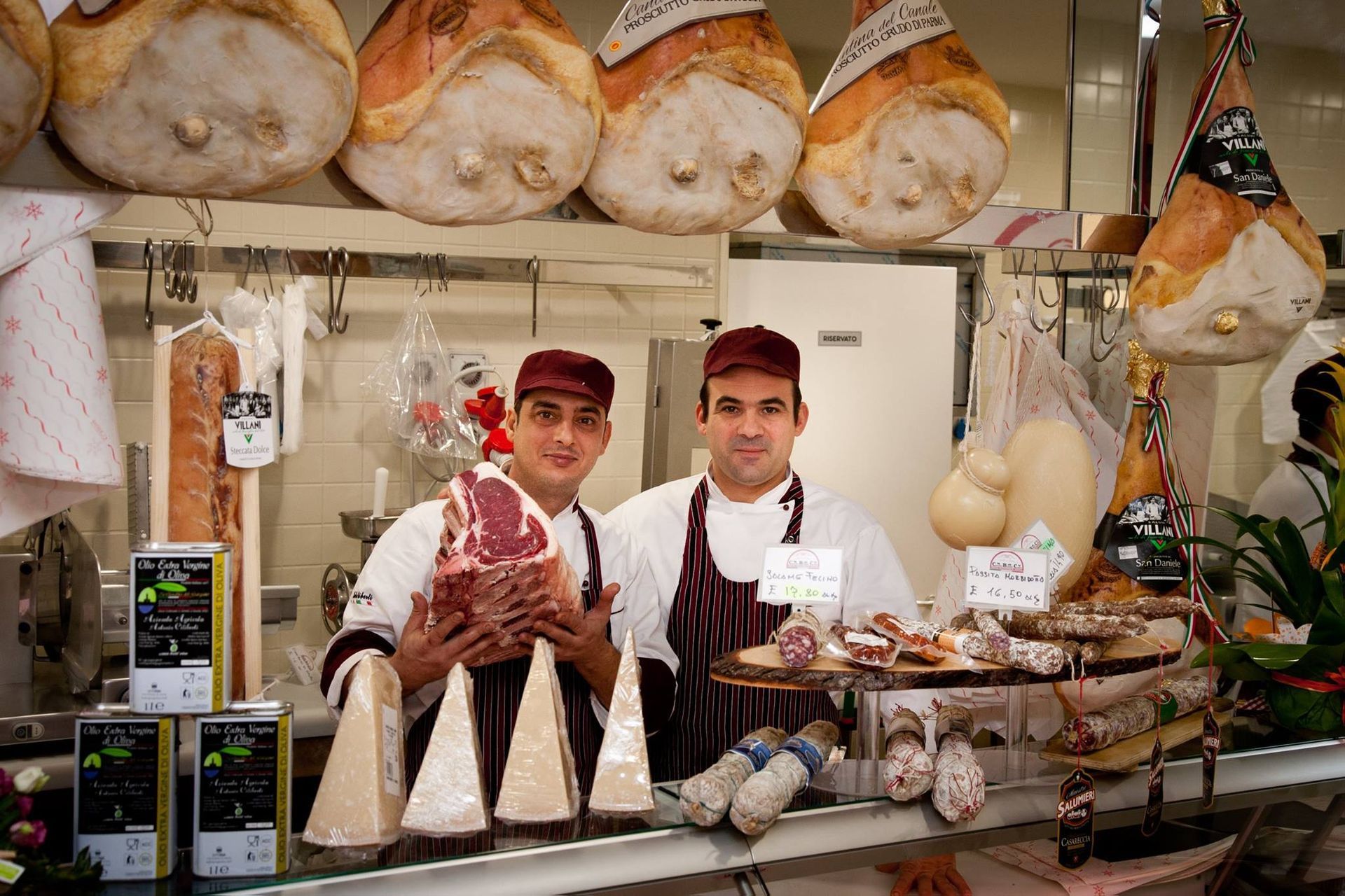Due uomini al bancone di una gastronomia con salumi e formaggi, tengono in mano un grosso prodotto a base di carne.