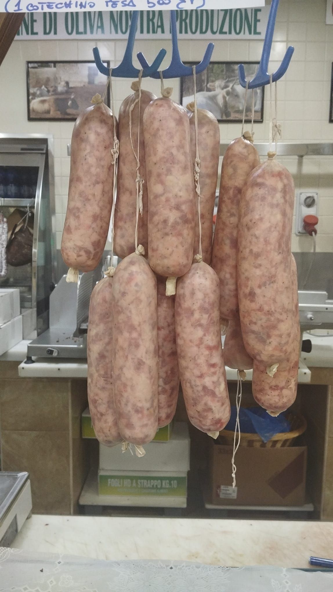 Salsicce appese a una rastrelliera blu in una macelleria. Sullo sfondo, bancone con bilance e scatole.