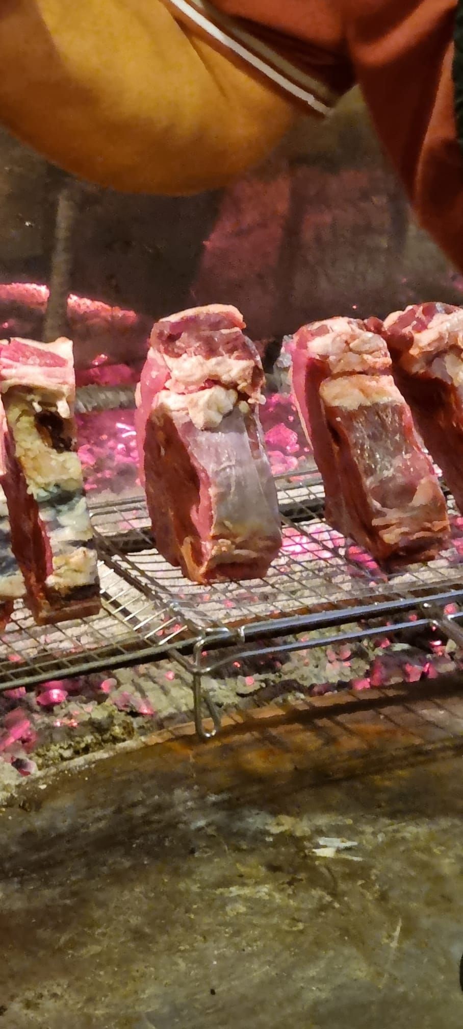 Bistecche cotte su una griglia metallica sopra braci ardenti. Sopra si vede il braccio di una persona.