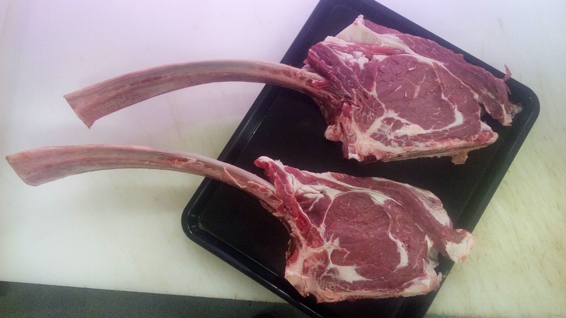 Due bistecche tomahawk crude su un vassoio nero, appoggiate su una superficie bianca.