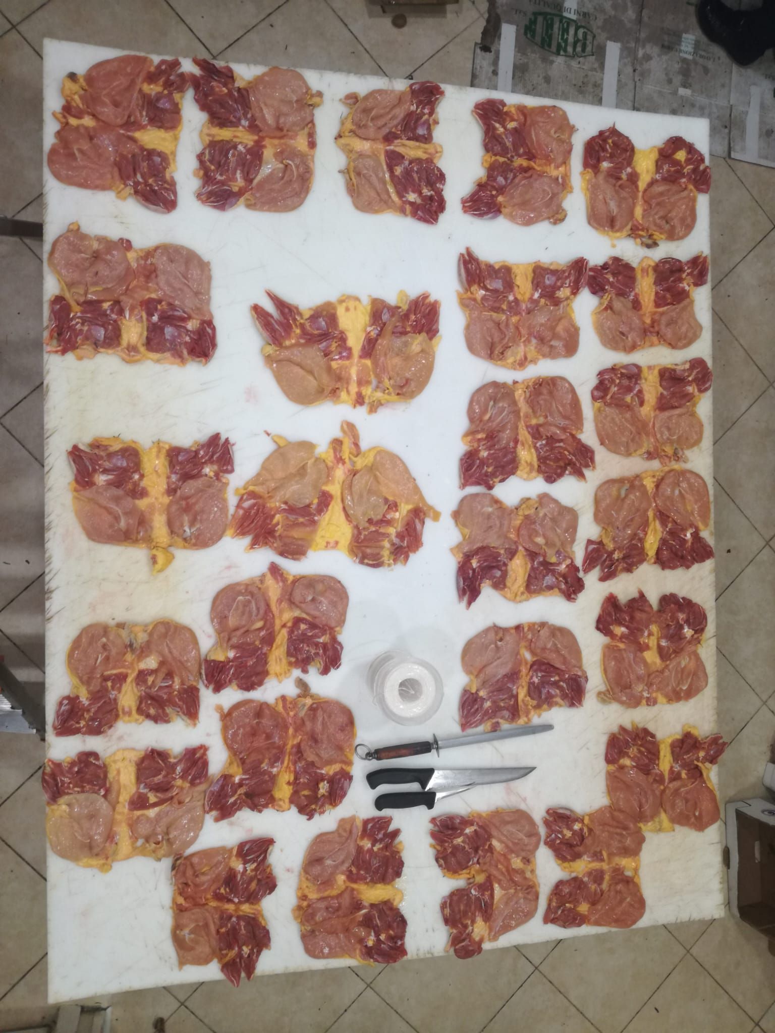 Porzioni di carne cruda disposte su un tavolo bianco con coltelli e nastro adesivo.