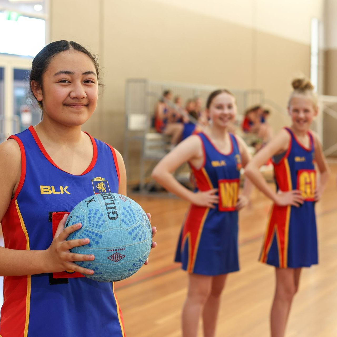 A Downlands student netballer in the foreground is holding a netball and there are two Downlands netballers in the background with their hands on their hips.