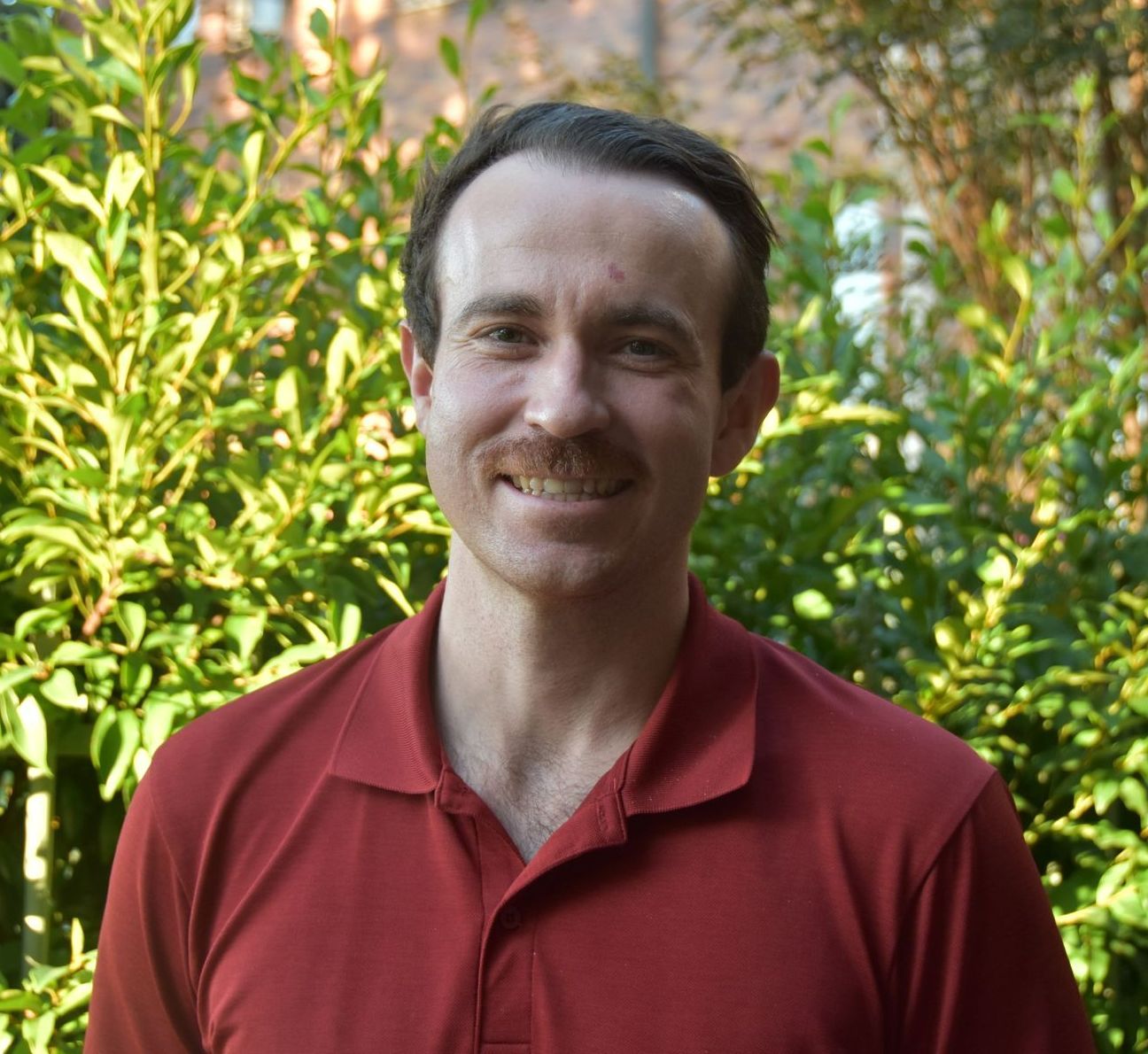 A man in a red shirt is smiling in front of a bush.