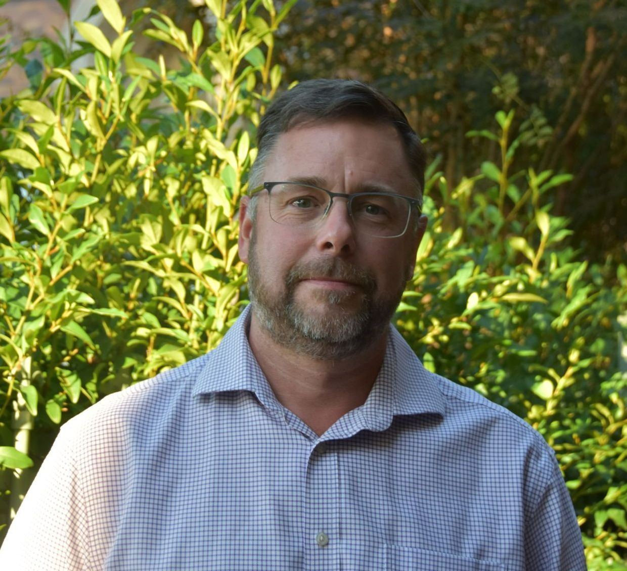 A man with glasses and a beard is standing in front of a bush.