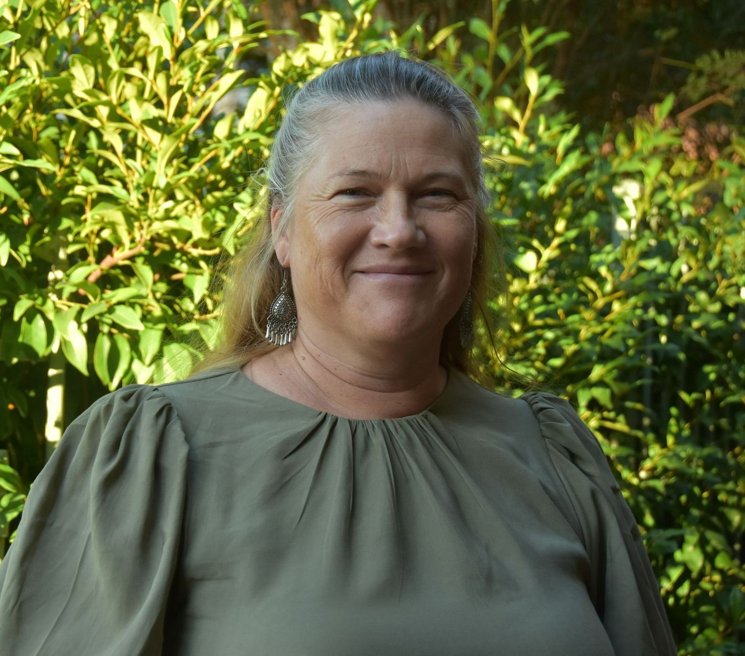 A woman in a green shirt is smiling in front of a bush
